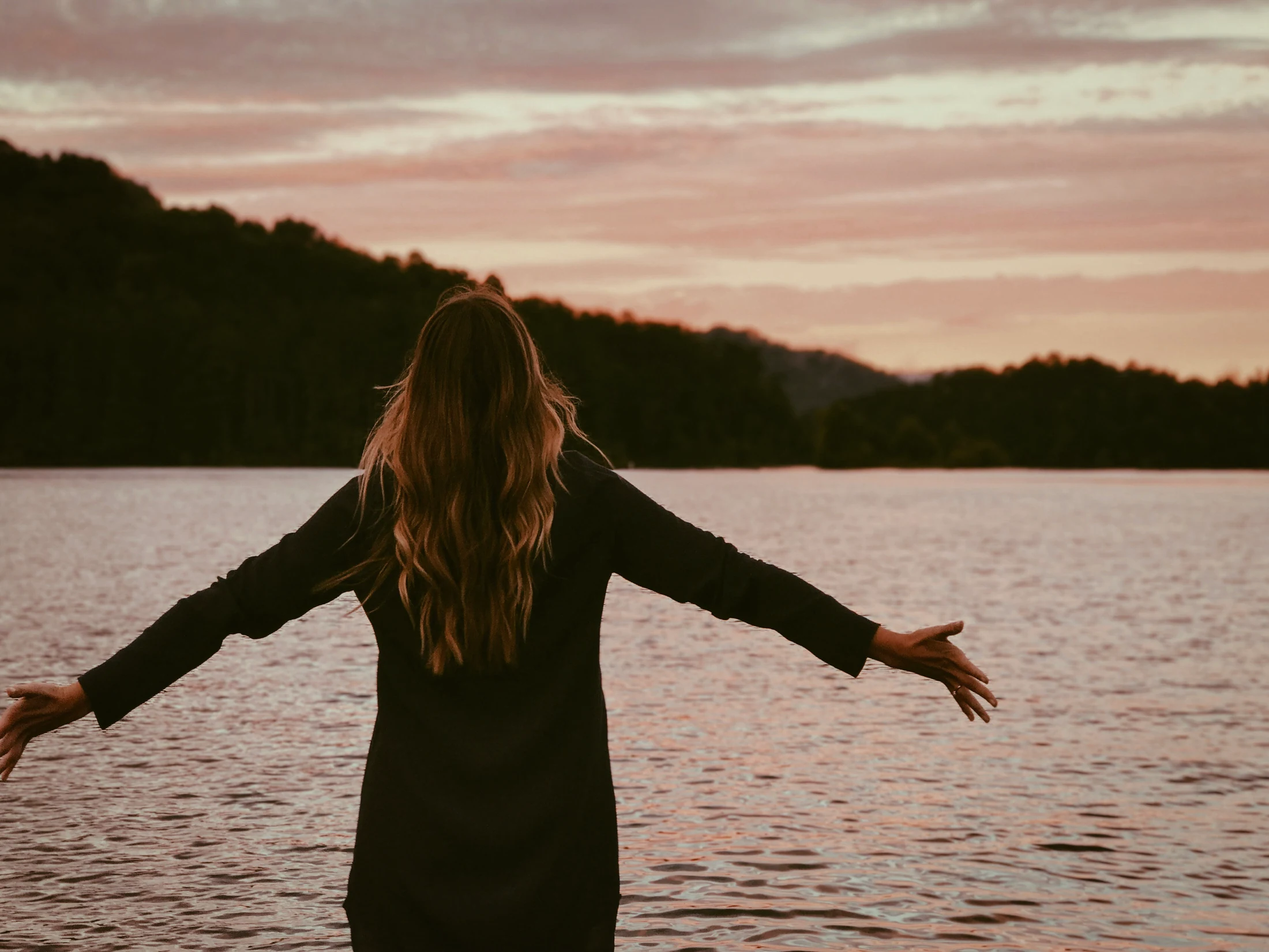 A person opens their arms facing the water at sunset